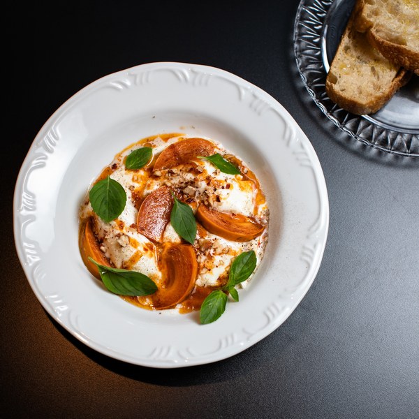 Burrata com caqui grelhado, manjericão e amêndoas, em prato branco.