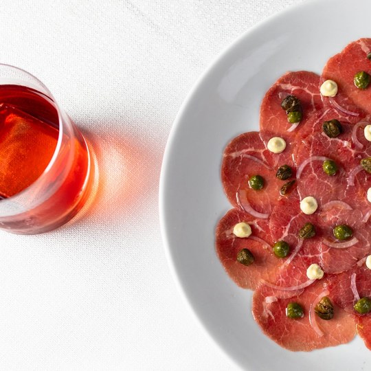 Negroni ao lado de um carpaccio di manzo sobre a mesa clara.