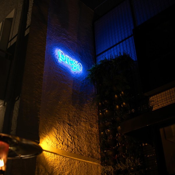Letreiro de neon azul com a assinatura Prego, aceso na parede de pedra da entrada.